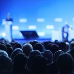 Crowd facing conference stage and speaker at podium